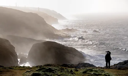 This landscape photograph, taken on a winter afternoon, captures the rugged cliffs of the Cornwall coast in the United Kingdom. The main subject of the image is a person looking out over the dramatic, mist-covered Cornish cliffs as waves crash against the rocky shoreline. In the background, several tall chimneys—reminiscent of Cornwall's historical mining heritage—rise above the cliffs, adding a distinctive feature to the scene. The photograph showcases the raw power of nature along this iconic coastline, with Gerald Weinbren’s composition emphasizing the interplay of light, sea spray, and rock formations.